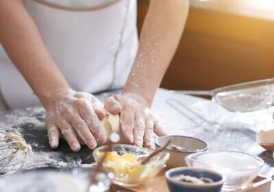 Hands of anonymous woman covered with flour kneading cookie dough on messy kitchen table, sunshine illuminating room