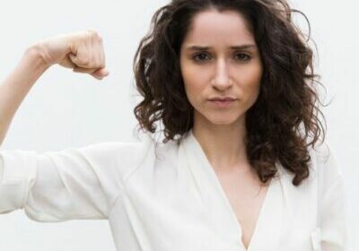 Confident serious woman flexing bicep, showing hand muscle. Wavy haired young woman in casual shirt standing isolated over white background. Girl power or feminism concept