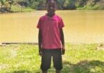 Menino Luiz Guilherme, de apenas 8 anos, vestindo bermuda preta e camiseta rosa em frente a uma lagoa